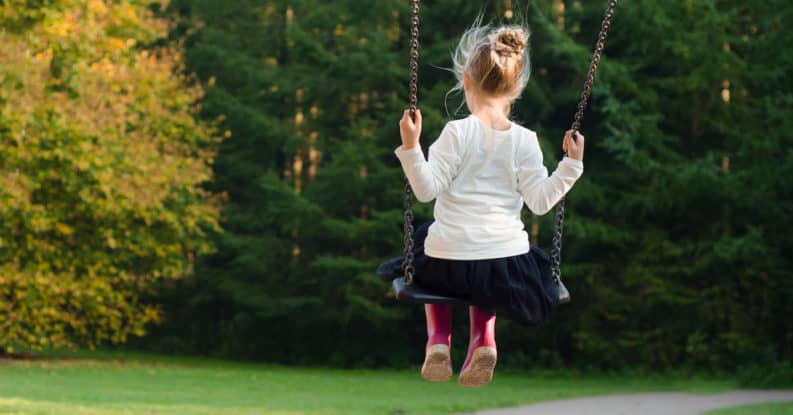 Young girl in a white shirt, black skirt and pink rain boots swinging on a park swing.